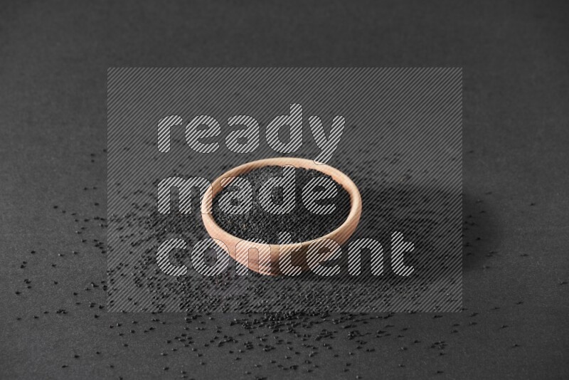 A wooden bowl full of black seeds surrounded by seeds on a black flooring