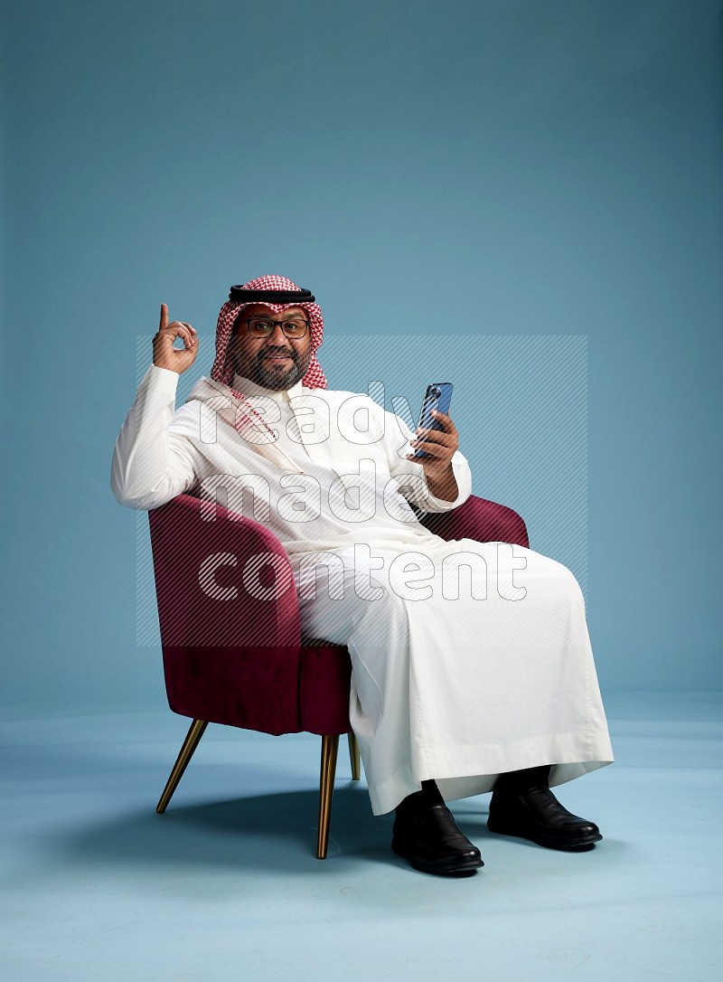 Saudi Man with shimag sitting on chair texting on phone on blue background