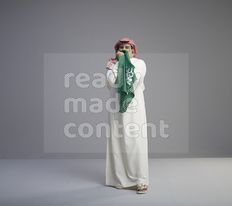 A Saudi man standing wearing thob and red shomag kissing big Saudi flag on gray background