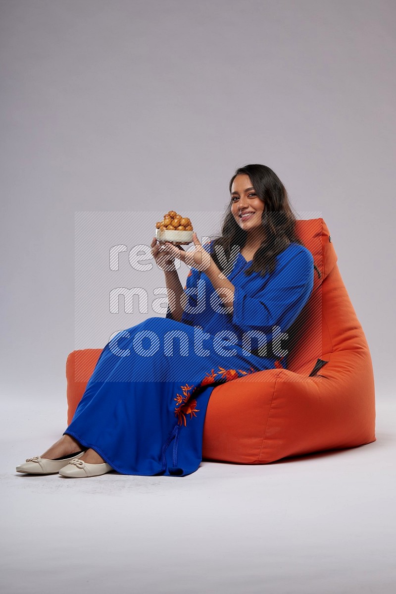 A Woman sitting on an orange beanbag wearing Jalabeya holding a plate of luqaimat