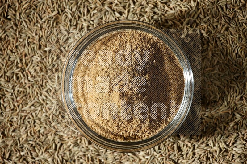 A glass bowl full of cumin powder surrounded by cumin seeds on black flooring