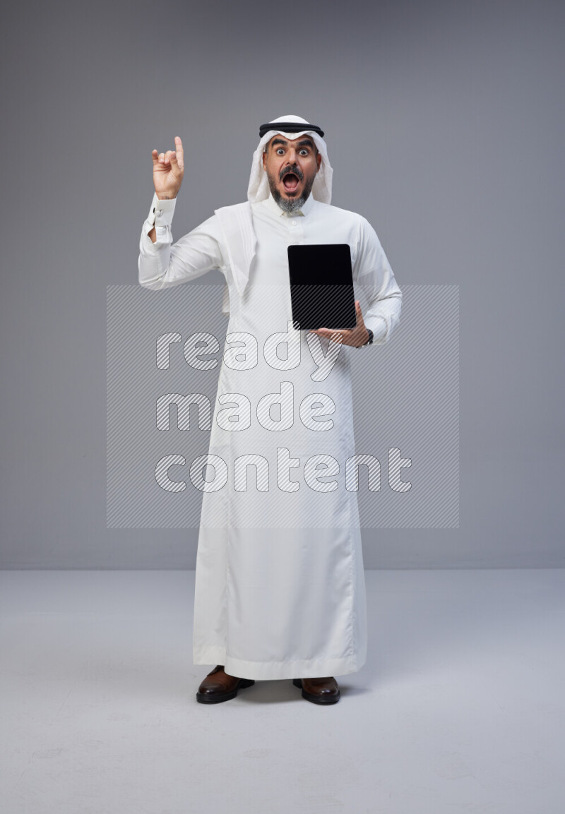 Saudi man Wearing Thob and white Shomag standing showing tablet to camera on Gray background