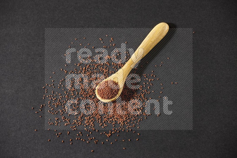 A wooden spoon full of garden cress seeds and seeds spread beside it on a black flooring