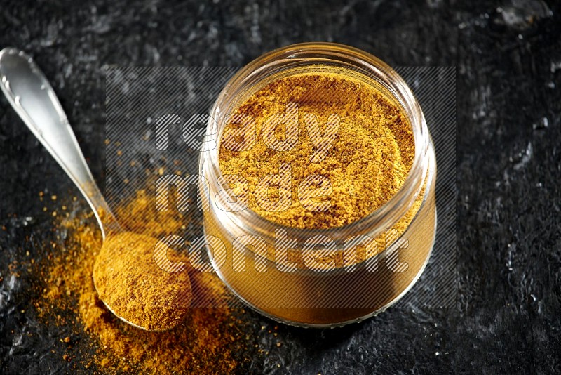 A glass jar and a metal spoon full of turmeric powder on a textured black flooring