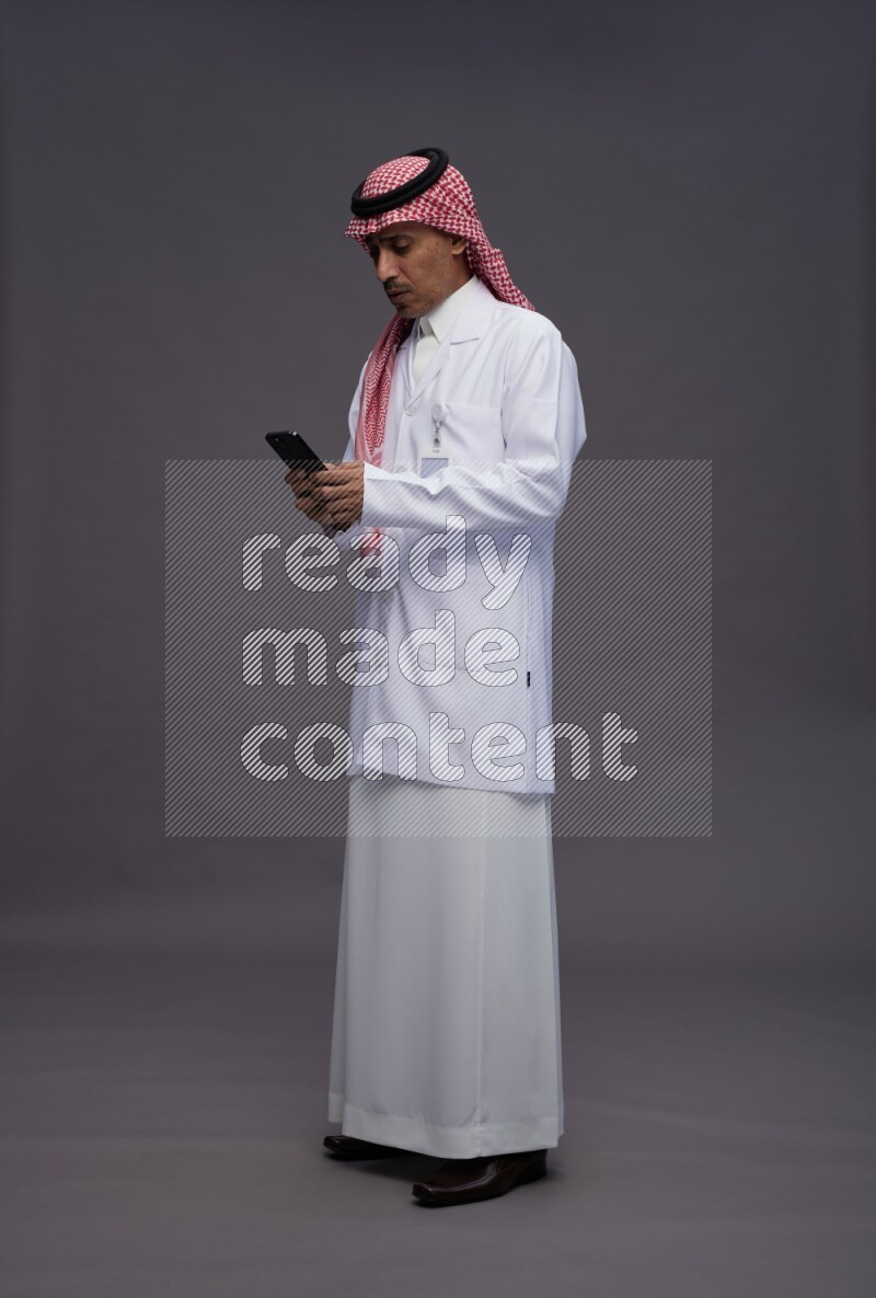 Saudi man wearing thob with lab coat and shomag with pocket employee badge standing texting on phone on gray background