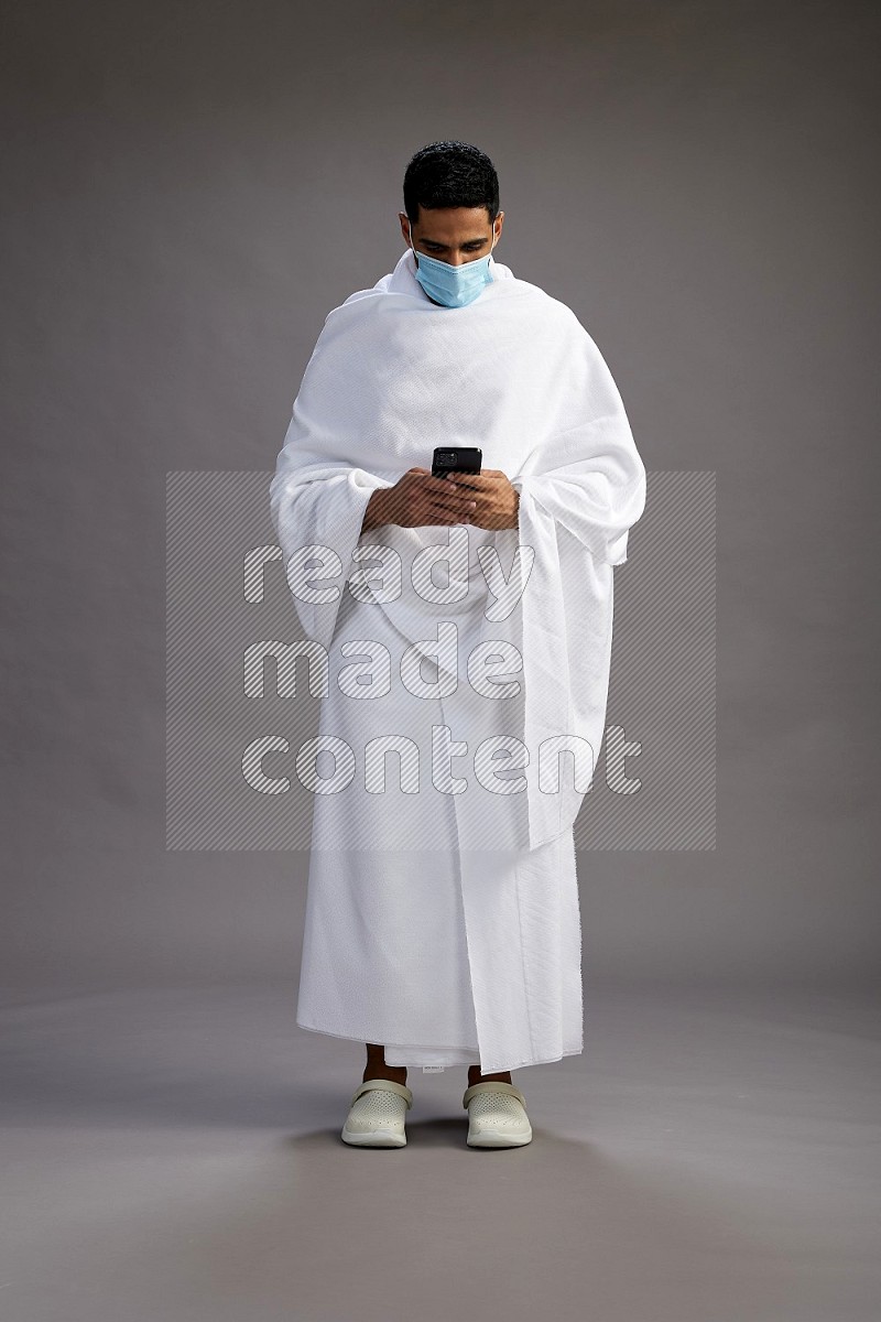 A man wearing Ehram with face mask Standing texting on phone on gray background