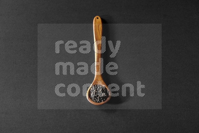 A wooden ladle full of black pepper beads on a black flooring