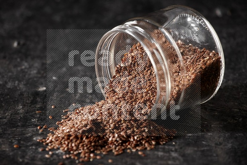 A glass jar full of flax flipped and seeds spreaded out on a textured black flooring in different angles