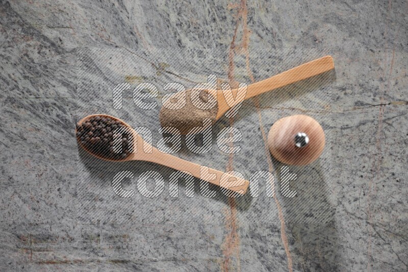 2 wooden spoons, one full of black pepper powder and the other full of black pepper beads and wooden grinder on a marble flooring