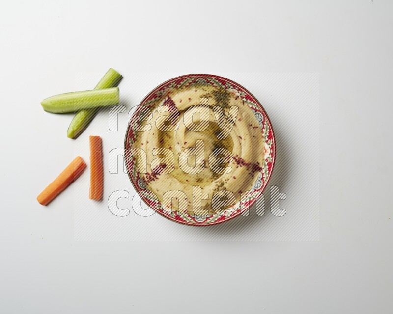 Hummus in a red plate with patterns garnished with zaatar & sumak on a white background
