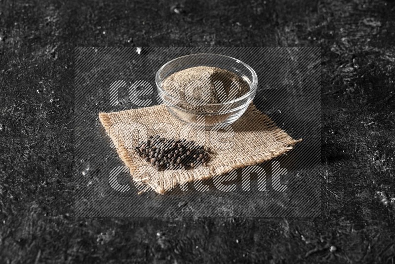 A glass bowl full of black pepper powder and black pepper beads on burlap fabric on textured black flooring
