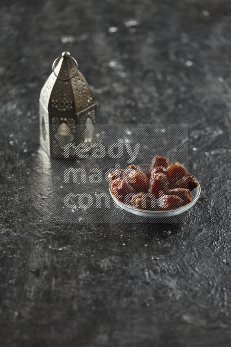 A silver lantern with different drinks, dates, nuts, prayer beads and quran on textured black background