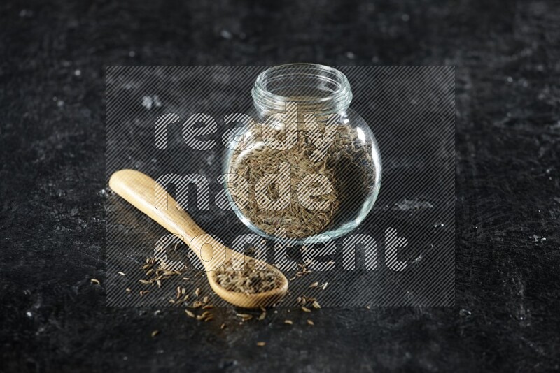 A glass spice jar and a wooden spoon full of cumin seeds on a textured black flooring