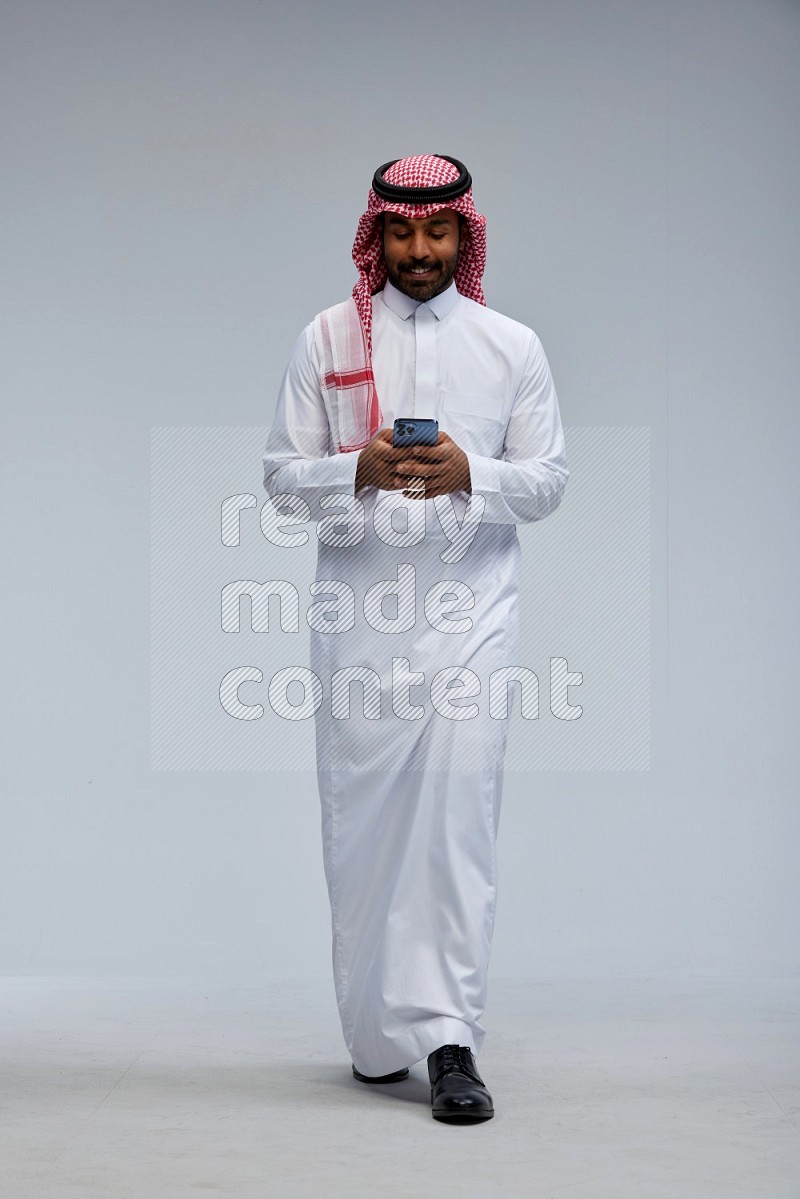 Saudi man Wearing Thob and shomag standing texting on phone on Gray background