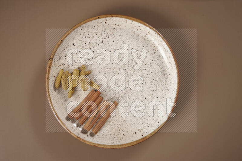 An assorted whole spices on a flat beige plate including fresh and dried ginger, cinnamon sticks, cardamom, turmeric fingers, cloves, star anise on a beige background