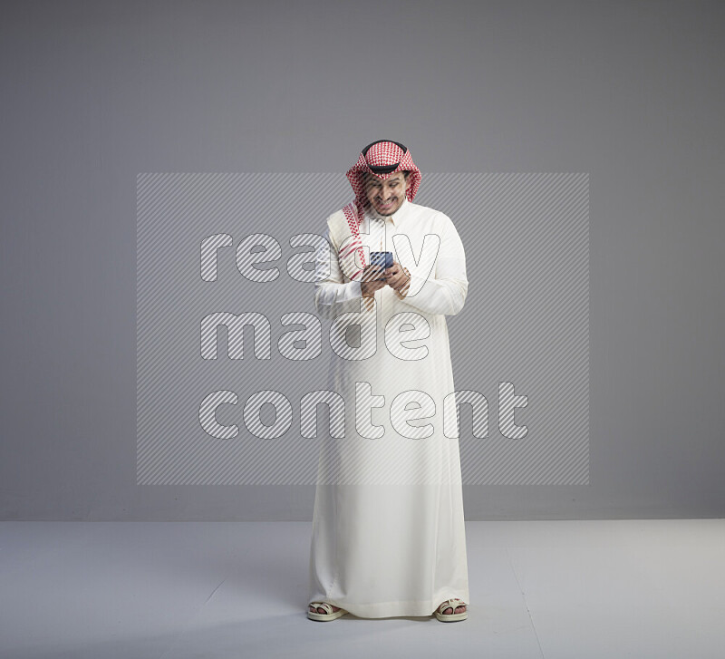 A  Saudi man standing wearing thob and red shomag texting on phone on gray background
