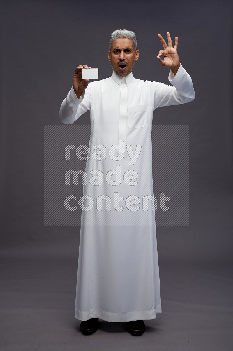 Saudi man wearing thob standing holding ATM card on gray background