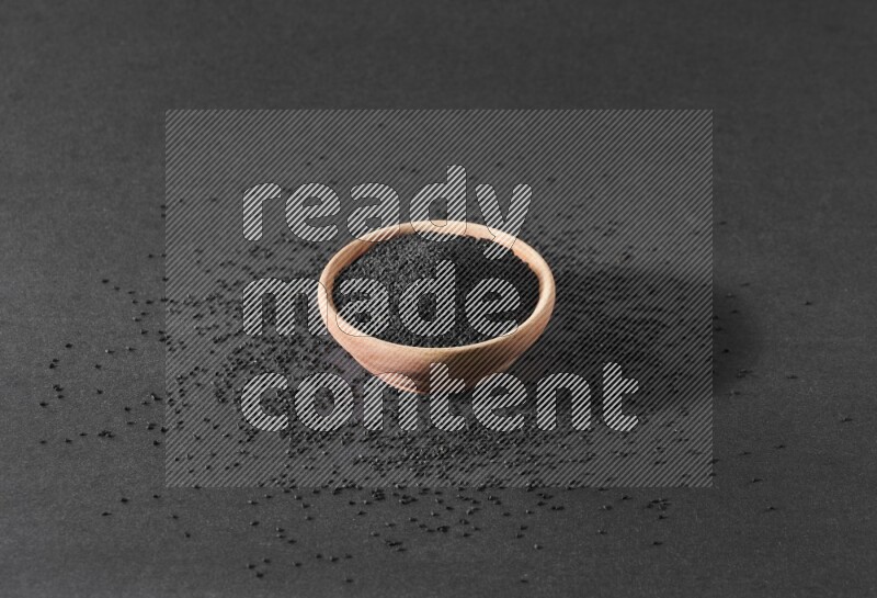 A wooden bowl full of black seeds surrounded by seeds on a black flooring