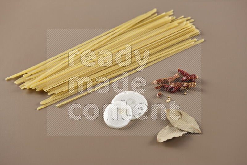 Raw pasta with different ingredients such as cherry tomatoes, garlic, onions, red chilis, black pepper, white pepper, bay laurel leaves, rosemary and cardamom on beige background
