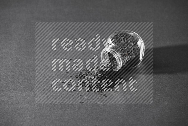 A flipped glass spice jar full of black seeds and the seeds spread out on a black flooring