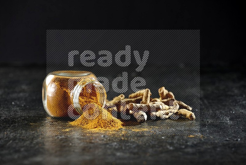 A flipped glass spice jar full of turmeric powder and powder spilled out of it with dried whole fingers on textured black flooring