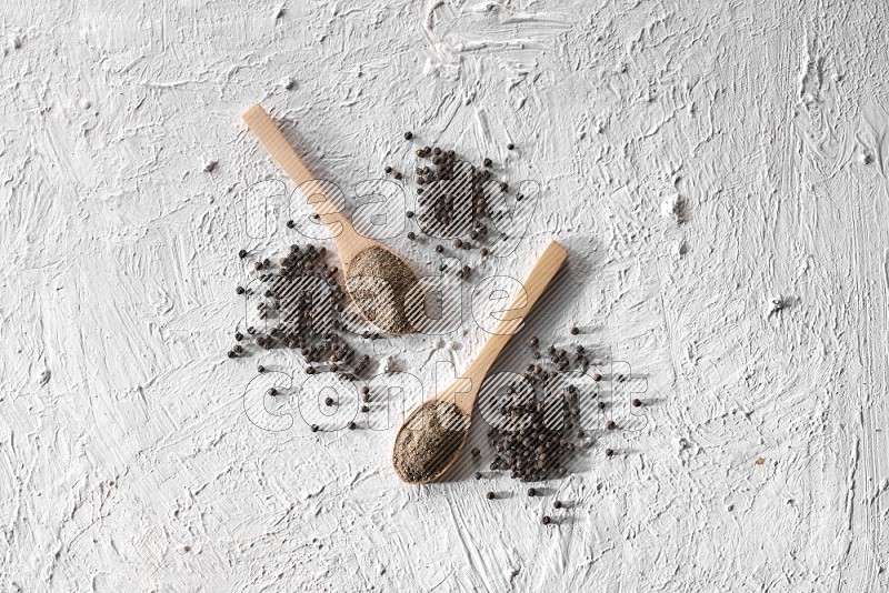 2 wooden spoons full of black pepper powder and black pepper beads spread on textured white background