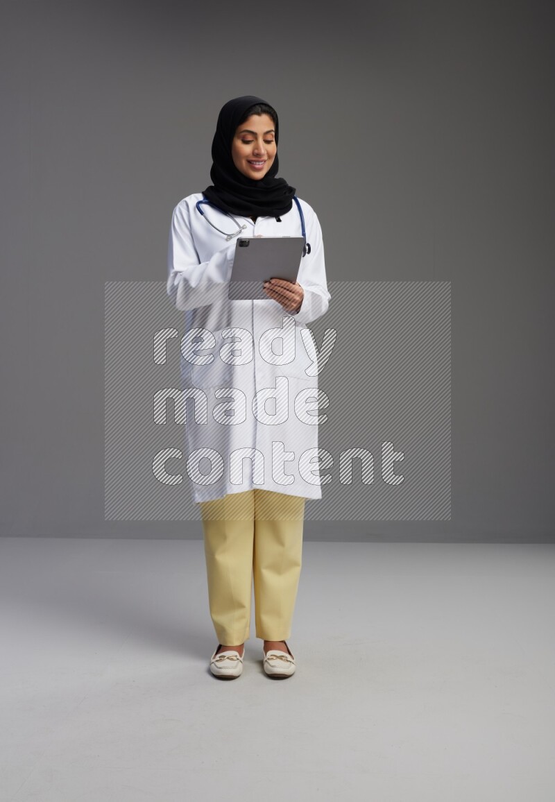 Saudi woman wearing lab coat with stethoscope standing working on tablet on Gray background