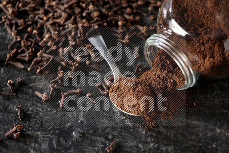 A flipped glass spice jar and a metal spoon full of cloves powder with cloves spread on textured black flooring