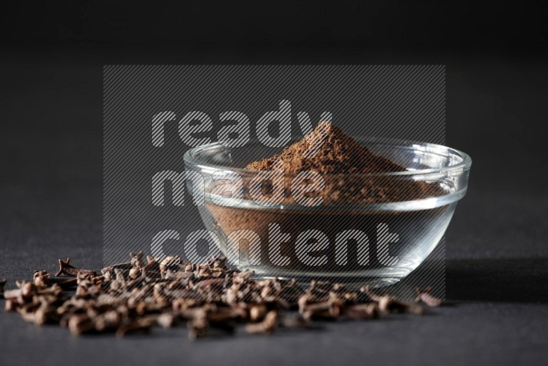A glass bowl full of cloves powder with cloves grains spread on black flooring