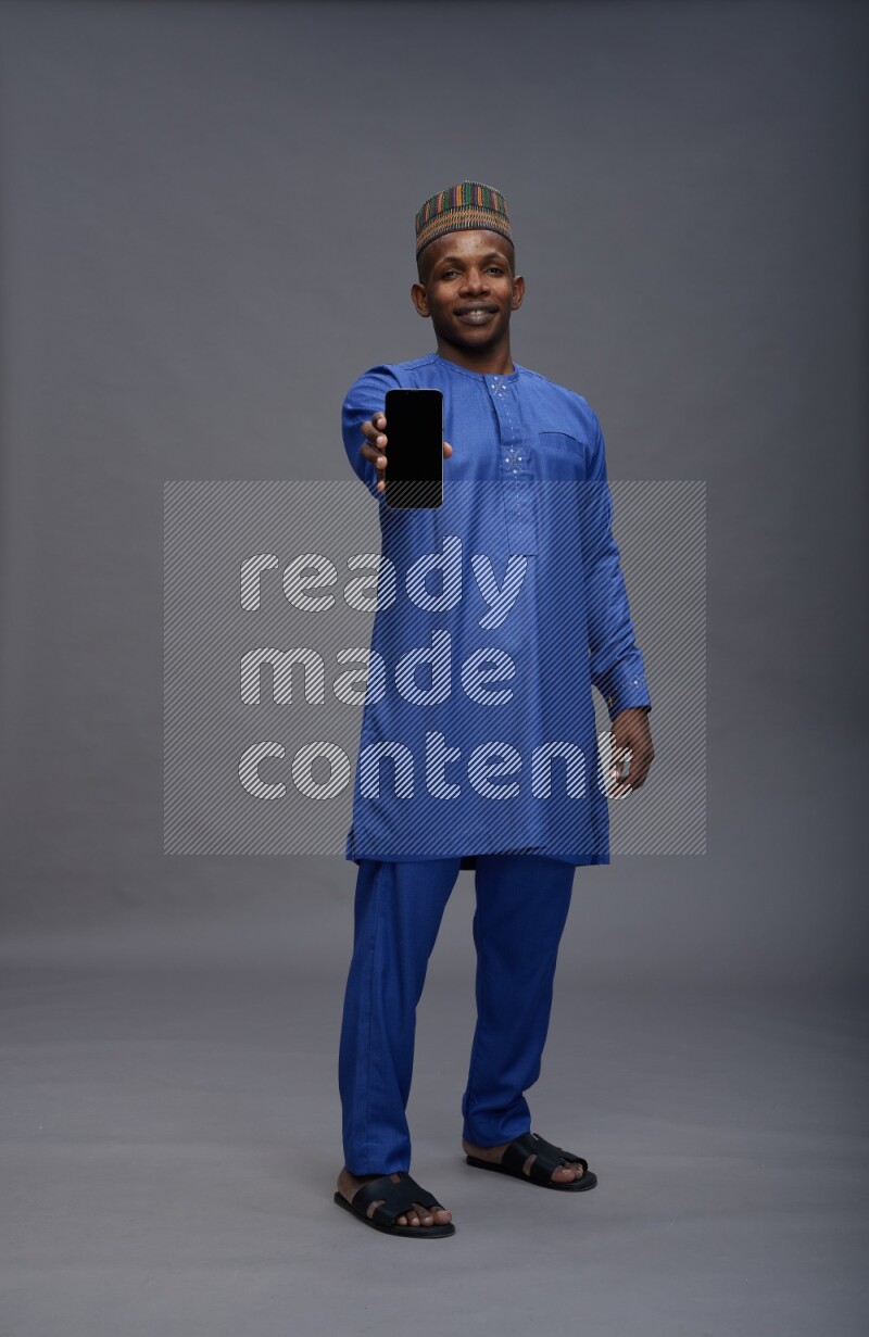 Man wearing Nigerian outfit standing showing phone to camera on gray background