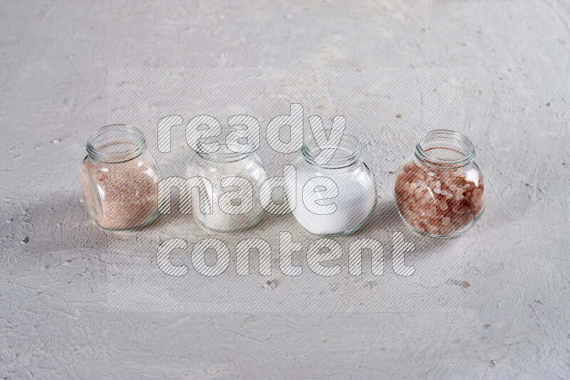 4 glass jars filled with table salt, coarse sea salt, fine himalayan salt and coarse himalayan salt on white background