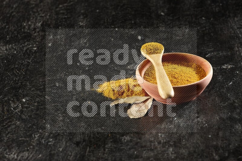 A wooden bowl and a wooden spoon full of turmeric powder on textured black flooring