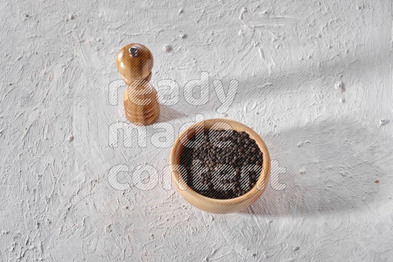 A wooden bowl full of black pepper and a wooden grinder on a textured white flooring