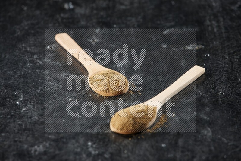 2 wooden spoons full of cumin powder on textured black flooring