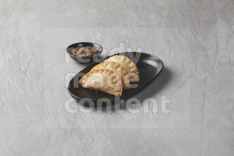 Three fried sambosas in an oval shaped black plate and a sauce in a black round ramekin on a gray background