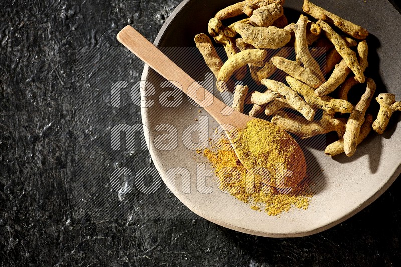 A plate filled with dried turmeric fingers and a wooden spoon full of turmeric powder on a textured black flooring