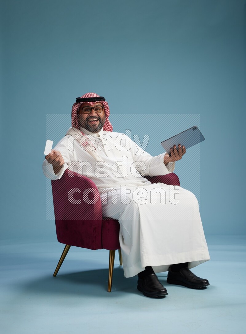 Saudi Man with shimag sitting on chair holding ATM card while working on tablet on blue background