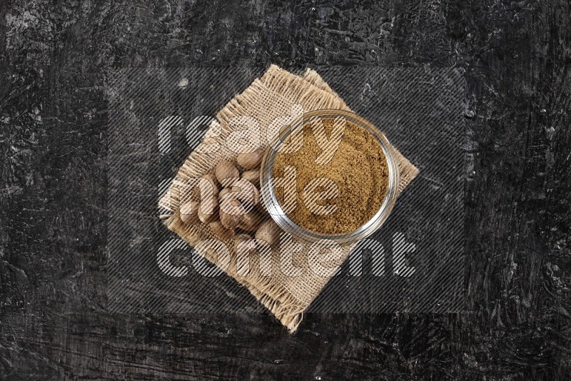 A glass bowl full of nutmeg powder with the seeds beside it on burlap fabric on a textured black flooring in different angles