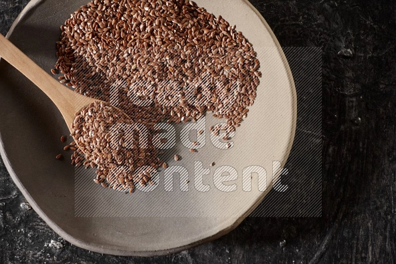 A multicolored pottery plate full of flaxseeds and wooden spoon full of seeds on a textured black flooring