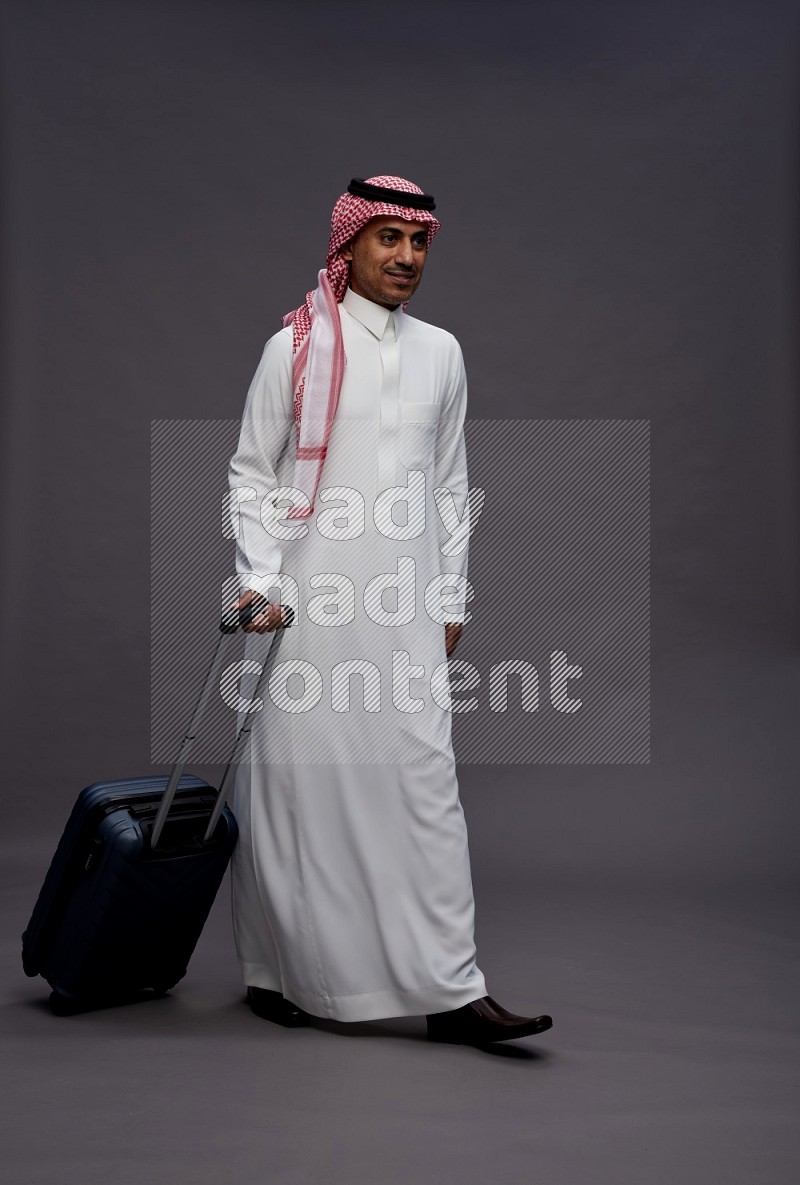 Saudi man wearing thob and shomag standing holding bag on gray background