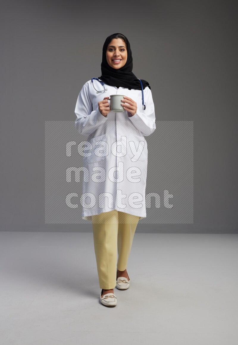 Saudi woman wearing lab coat with stethoscope standing holding mug on Gray background