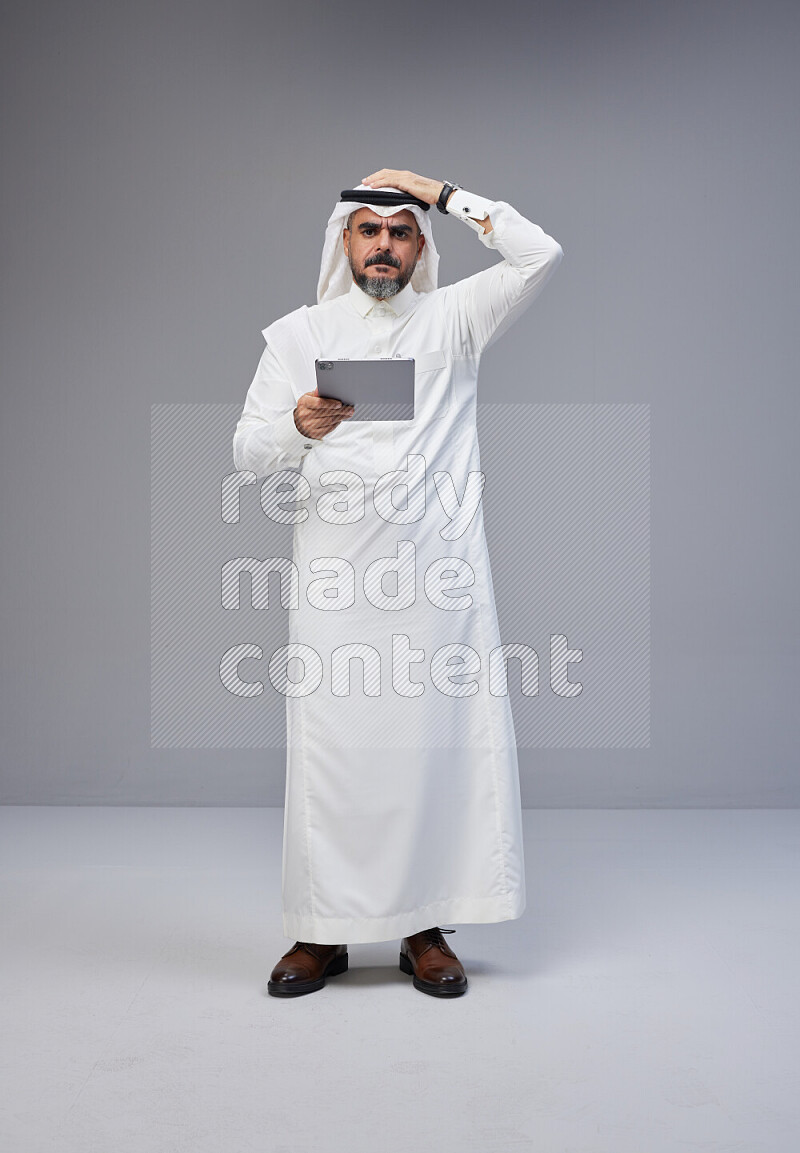 Saudi man Wearing Thob and white Shomag standing working on tablet on Gray background