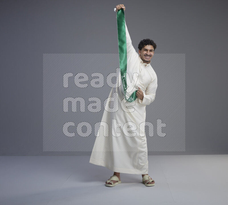 A Saudi man standing wearing thob holding Saudi flag scarf on gray background