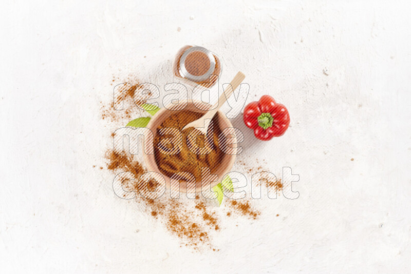 A wooden bowl full of ground paprika powder with a glass jar beside it and red bell pepper on white background