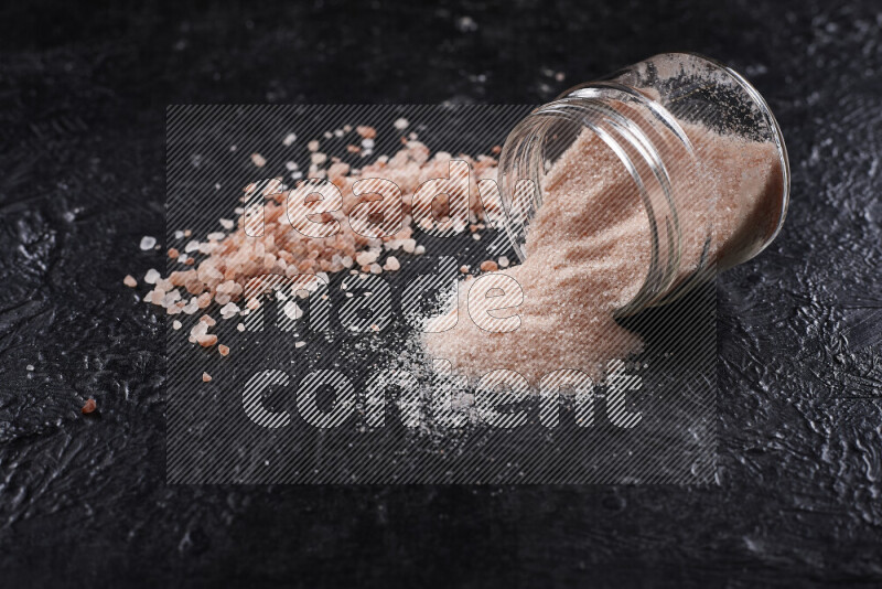 A glass jar full of fine himalayan salt with some himalayan crystals beside it on a black background