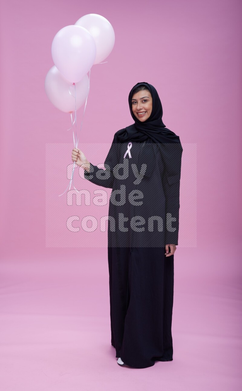Saudi woman wearing pink ribbon on Abaya standing holding balloons on plain pink background