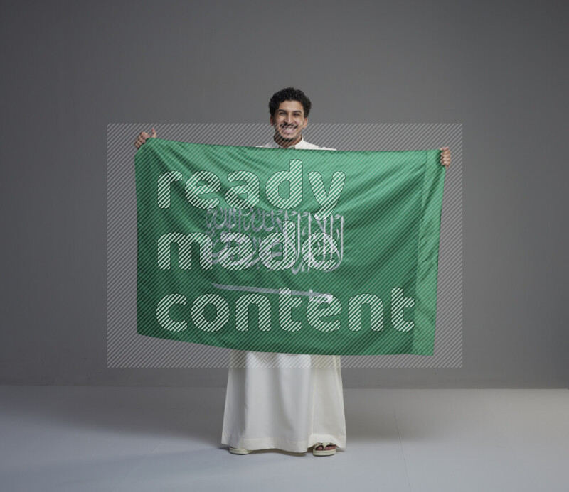 A Saudi man standing wearing thob holding big Saudi flag on gray background
