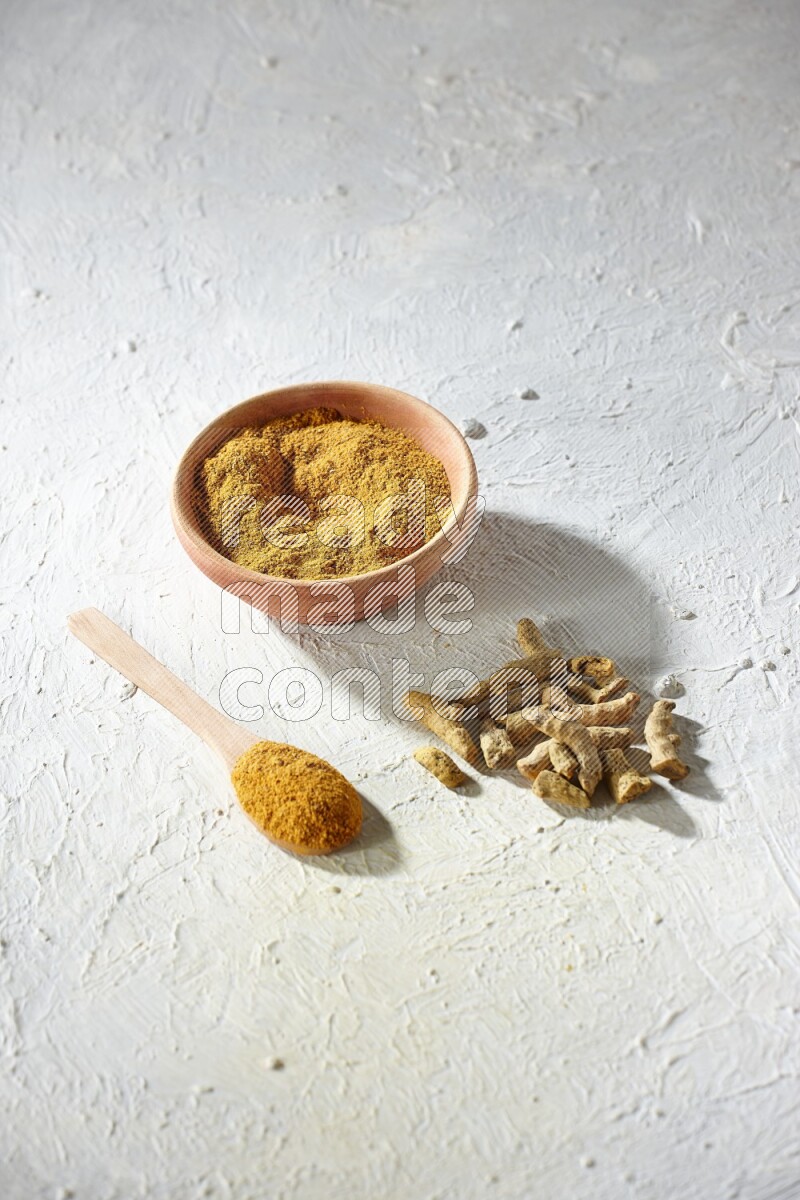 A wooden bowl and wooden spoon full of turmeric powder with dried turmeric fingers on textured white flooring