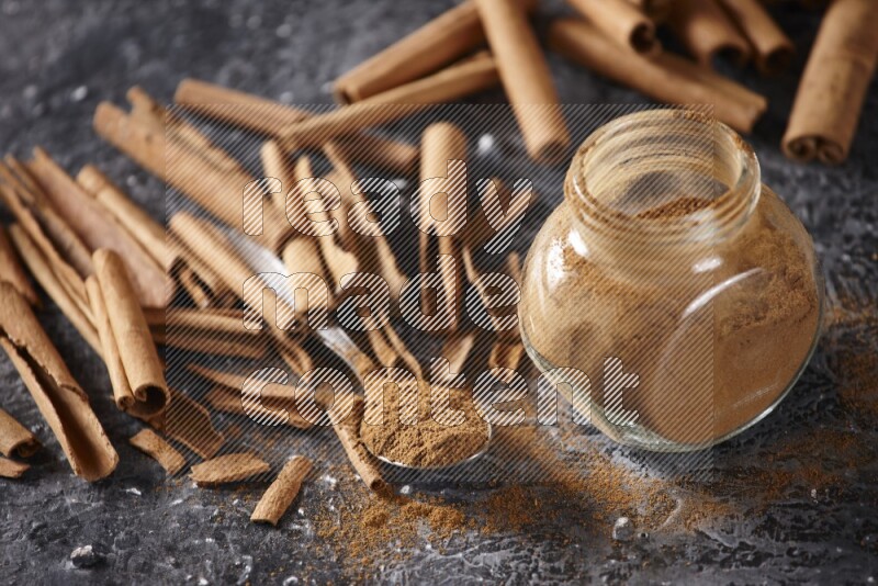Herbal glass jar and a metal spoon full of cinnamon powder surrounded by cinnamon sticks on textured black background