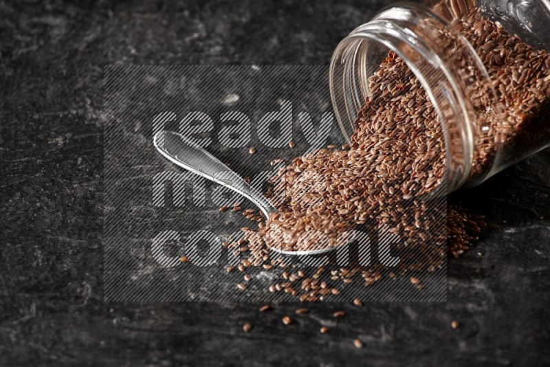 A glass jar full of flaxseeds flipped and seeds spread out with a metal spoon full of the seeds on a textured black flooring